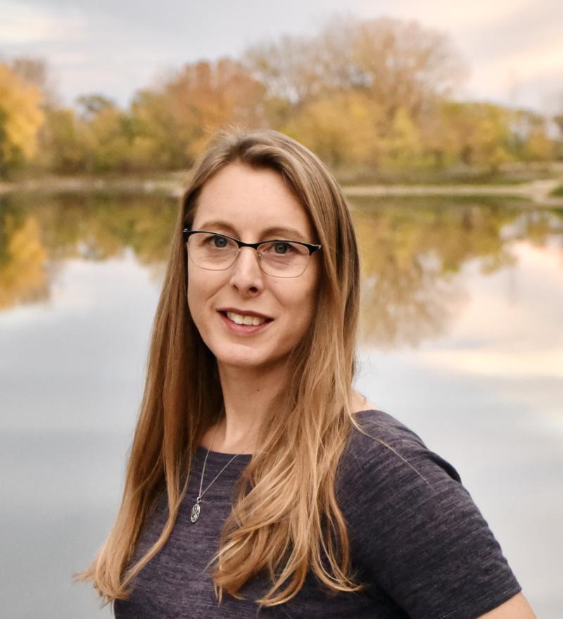 Robin Buterbaugh smiles at the camera while standing in front of a body of water with trees in the distant background