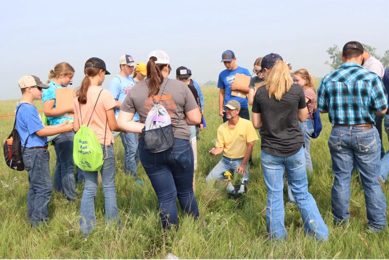 A man in a yellow shirt kneels in a pasture with a group of youth and teenagers standing in a circle around him