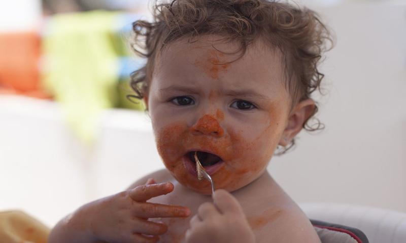 Infant grasping a training fork with tomato sauce all over her face.
