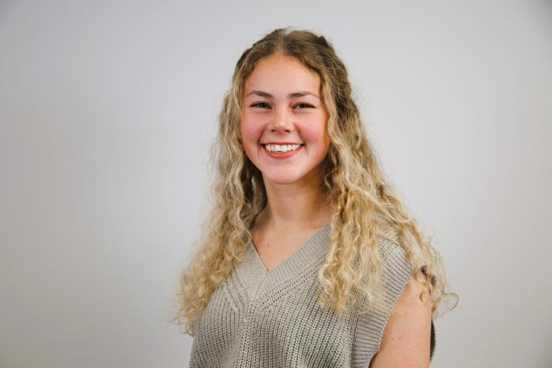 Addie Womack smiles at the camera. She has long, curly blonde hair and is wearing a grey shirt