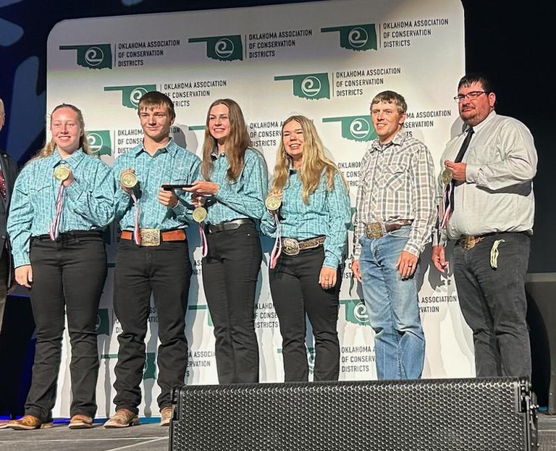 A team of four people in matching blue button-down shirts stands on stage with their awards, with two people off to the right side