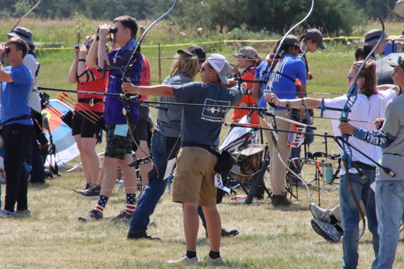 Youth are lined up taking aim with bows and arrows to shoot at outdoor targets