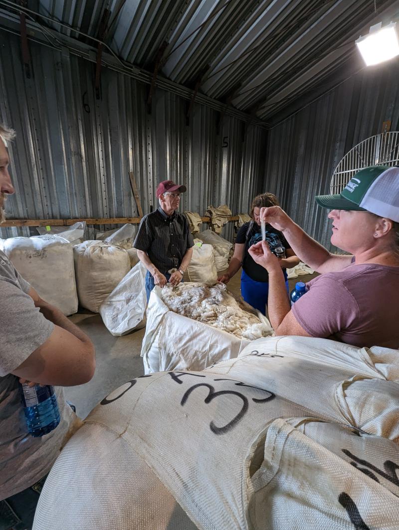 A man in a warehouse surrounded by bags of wool speaks to a group of people