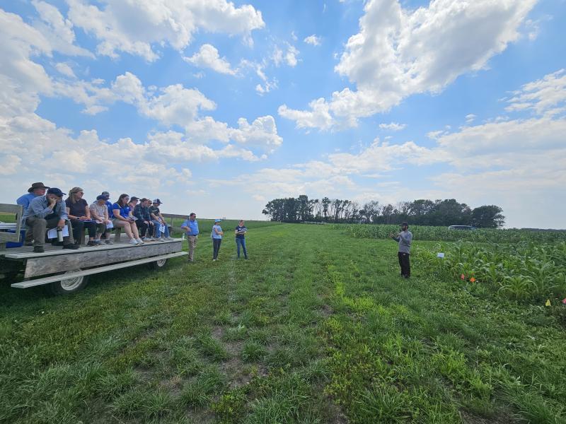 A group of people sits on an open trailer and listens to a man giving a presentation in front of a field