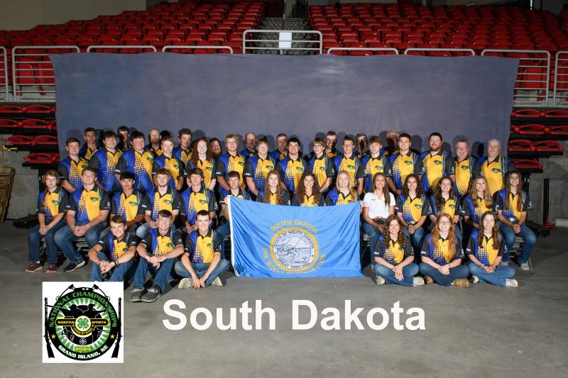 A large group of youth in matching blue and yellow shirts pose. They are holding a South Dakota flag in front of them