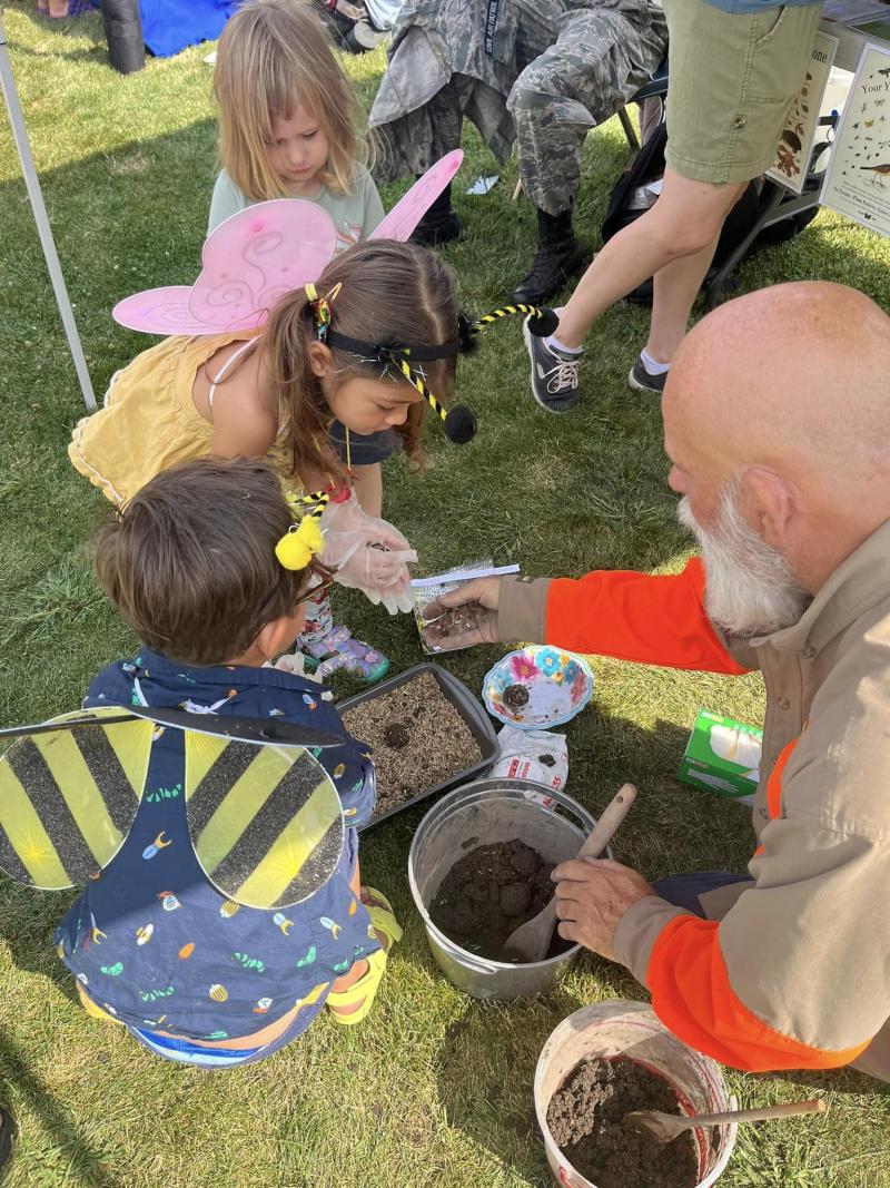 A little girl wearing butterfly wings and a little boy wearing honeybee wings scoop dirt and insects out of pails