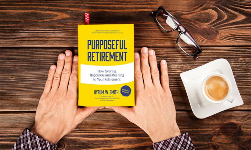 Hands holding a copy of the book, “Purposeful Retirement: How to Bring Happiness And Meaning to Your Retirement” at a wood table with a cup of coffee and a pair of reading glasses nearby.