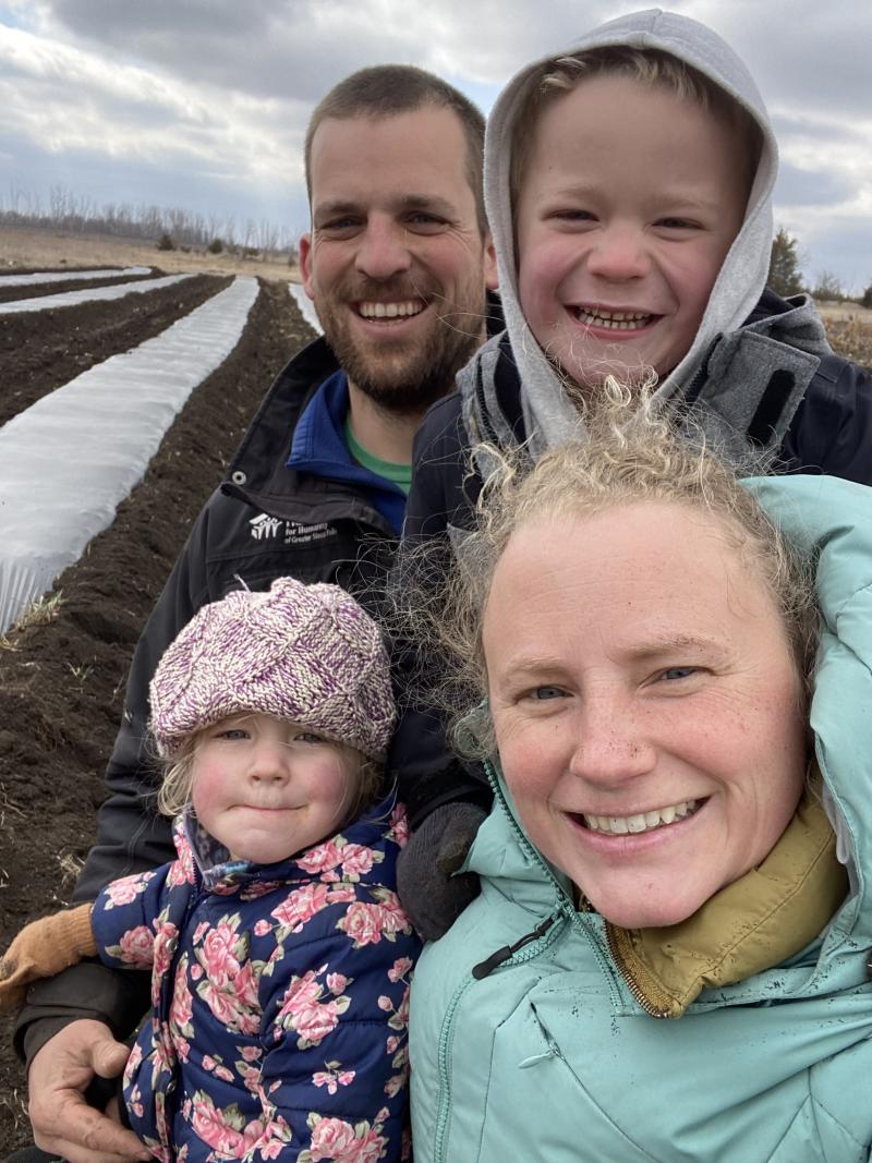A man, woman and two small children smile for a selfie in front of rows of vegetables
