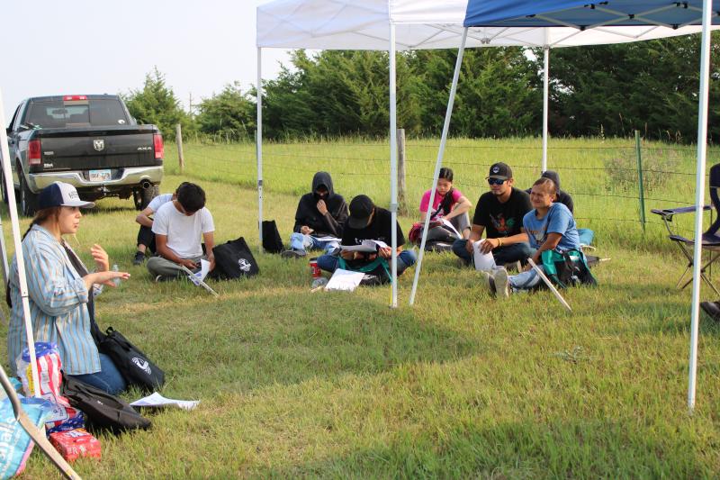 A woman in a baseball cap and long-sleeved shirt kneels on the ground while speaking to a group of people sitting in the grass in a pasture