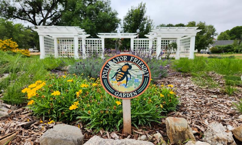 Sign with a picture of a bee and Pollinator Friendly Garden text, with flowers and a white gazebo in the background.