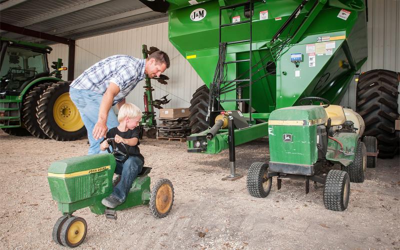 Father carefully watching his son on a toy tractor in a machine shed.