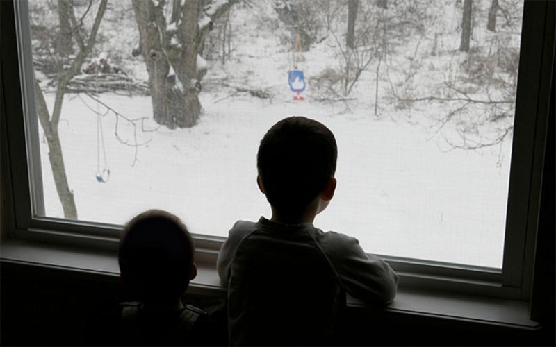 Two children look out their living room window at a snowy winter landscape.
