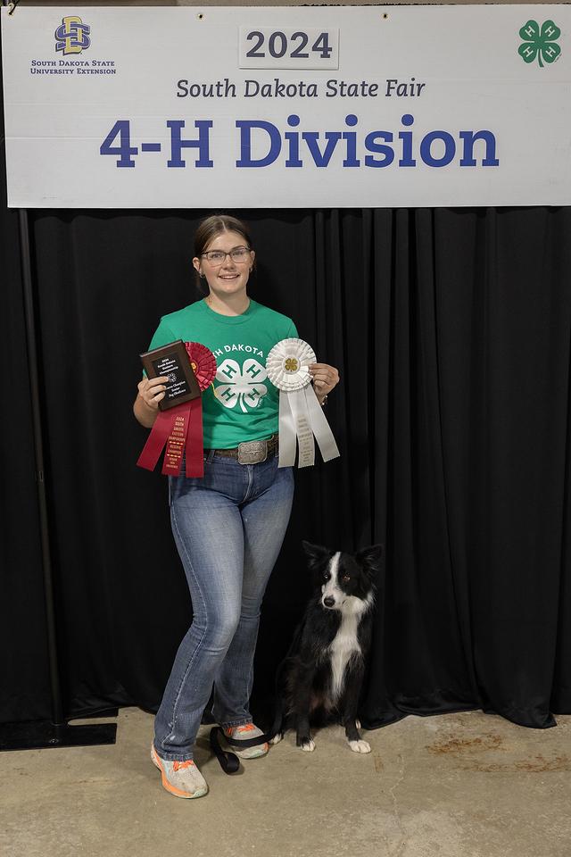 A girl in a green shirt holds up a plaque and large red and white rosettes; a black and white dog sits beside her