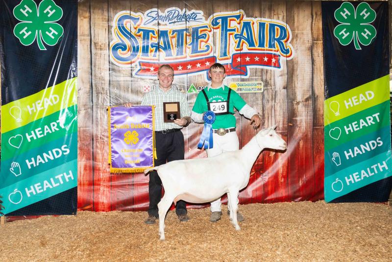 A teenage boy holds a goat by the collar and holds up a blue ribbon. Someone beside him is holding up a banner and a plaque