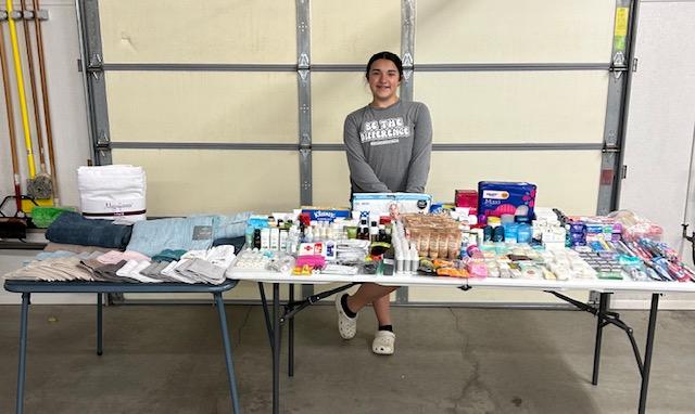 A girl stands behind two long tables stacked with donations of personal hygiene items