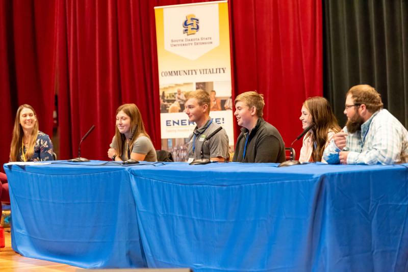 A panel of people sits behind tables on a stage with an Energize banner behind them