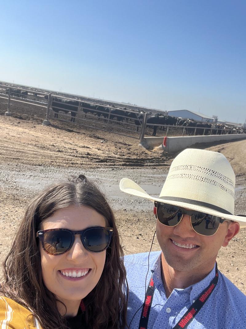 A man and a woman smile for a selfie in front of a feedlot