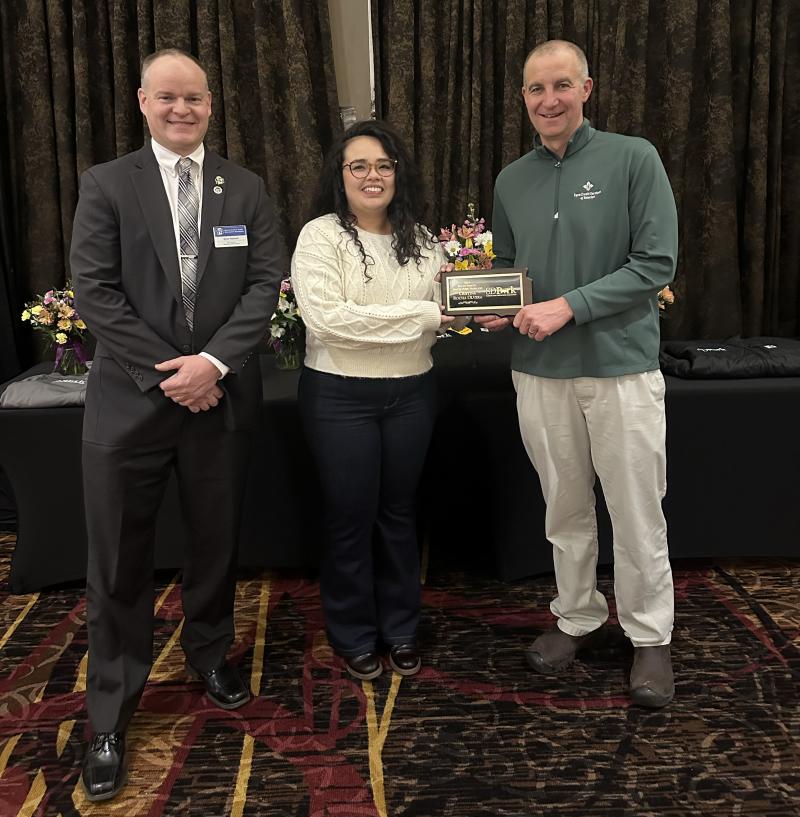 Three people smile for the camera. The woman in the center is holding an award plaque
