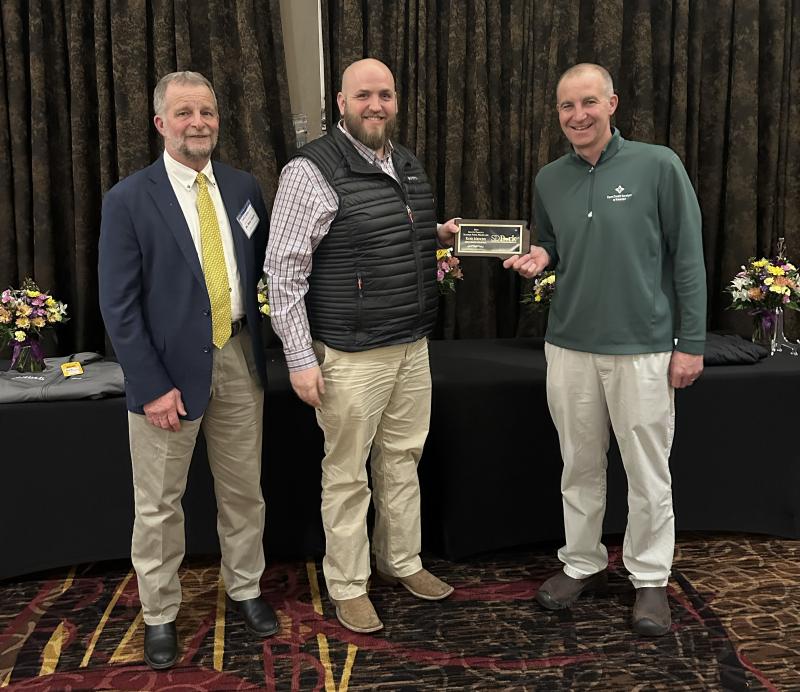 Three people smile for the camera. The man in the center holds a plaque.