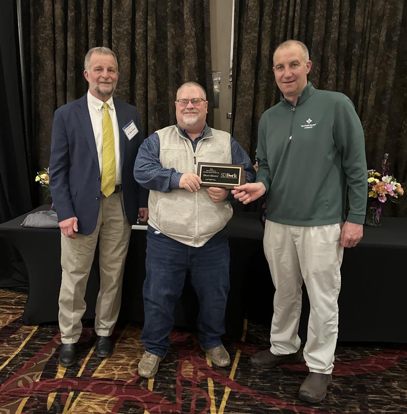 Three men smile for the camera. The man in the center holds a plaque
