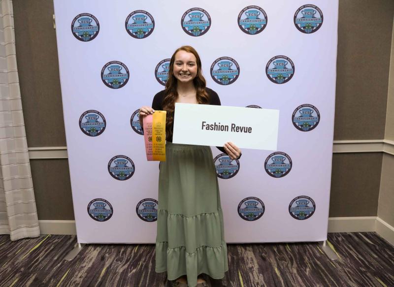Samantha Brenden smiles with yellow ribbons and a sign that says "Fashion Revue"