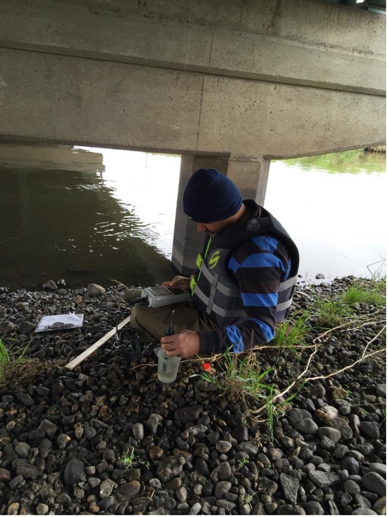 A man kneels next to a creek with testing equipment spread in front of him
