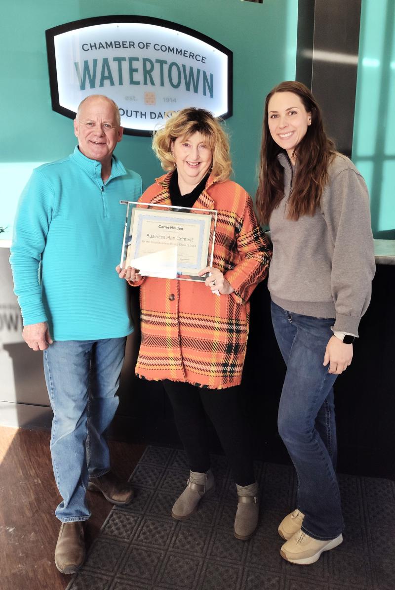 Three people smile for the camera; the woman in the middle holds up a certificate in a glass frame