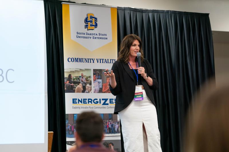 A woman in a black top and white skirt stands on a stage with a microphone