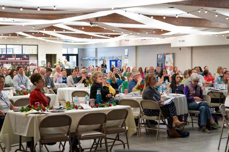 A large crowd of people sit at tables looking toward a speaker