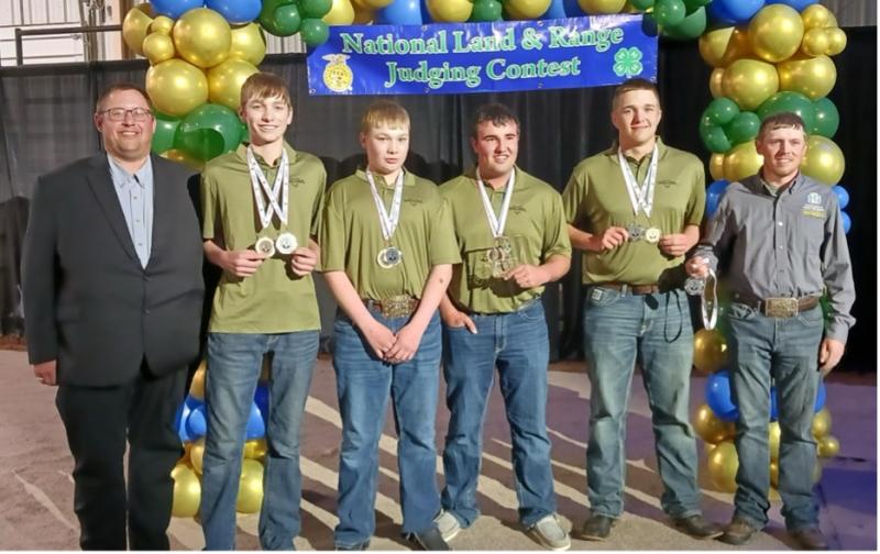 Four teenage boys in matching olive green shirts stand between their coaches wearing medals around their necks