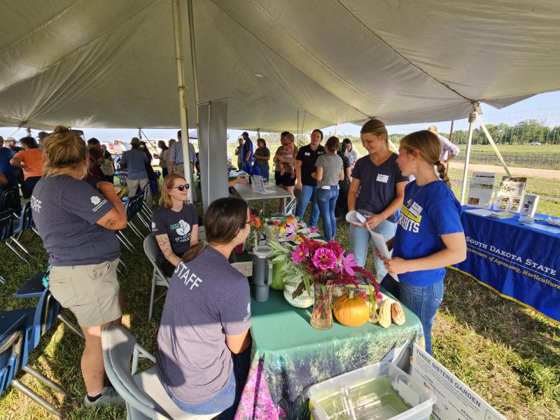 People gather around tables of produce and cut flowers under a tent