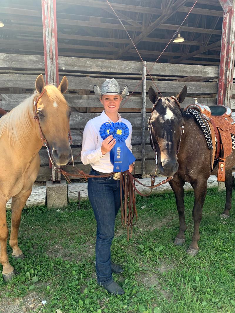 A girl in a cowboy hat stands between two horses and holds up two large blue rosettes