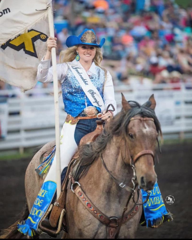 A woman wearing a cowboy hat with a crown rides a horse and carries a flag