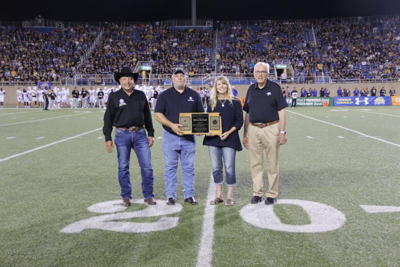 Four people stand on the 20-yard line of a football field. There are two men on the far left and far right. In the middle is a man handing a woman a wooden plaque in the shape of South Dakota.