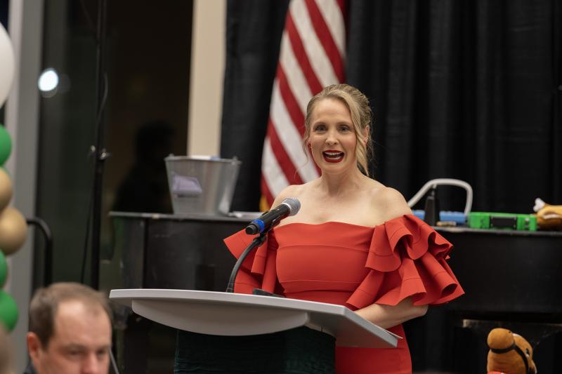 A woman with blonde hair and wearing a red dress speaks behind a podium
