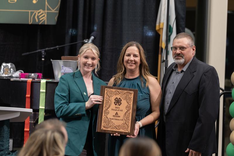 Three people hold up a wooden "Friends of 4-H" award plaque