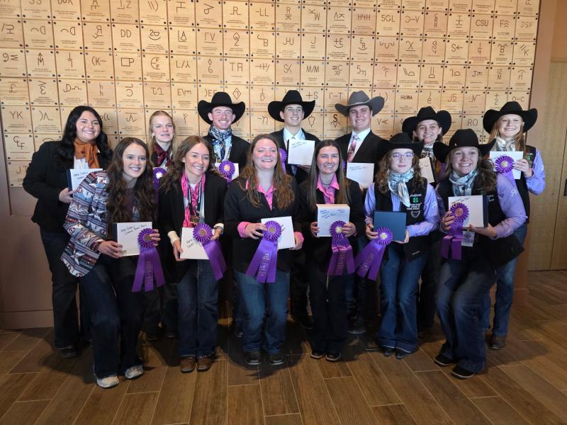 Thirteen youth in cowboy hats and western wear smile for the camera, holding up their ribbons and award plaques