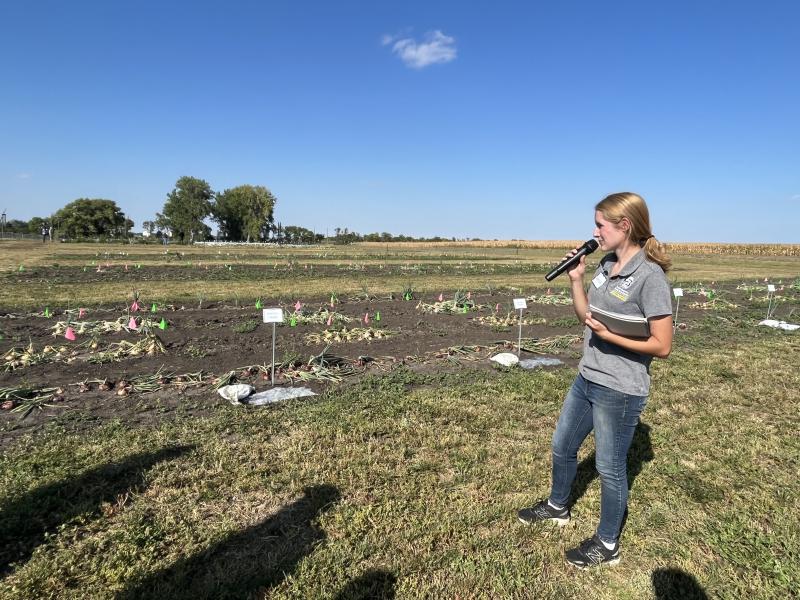A girl with a blonde ponytail, wearing a grey T-shirt with the SDSU logo and jeans holds a microphone standing next to vegetable plots
