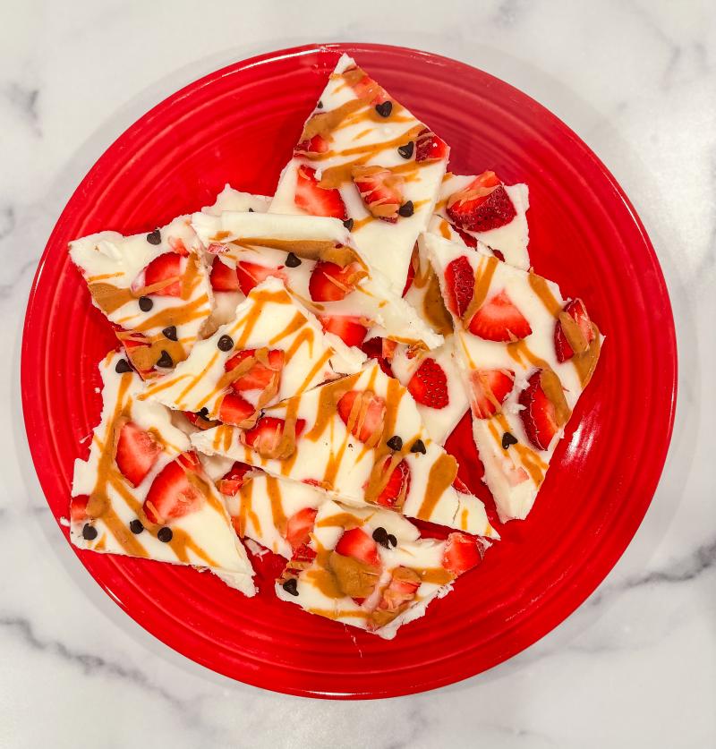 vanilla frozen yogurt bark with strawberries, blueberries and peanut butter drizzle in a red bowl on a kitchen counter.