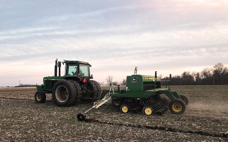 A green tractor pulls a planter to plant oats in a field.