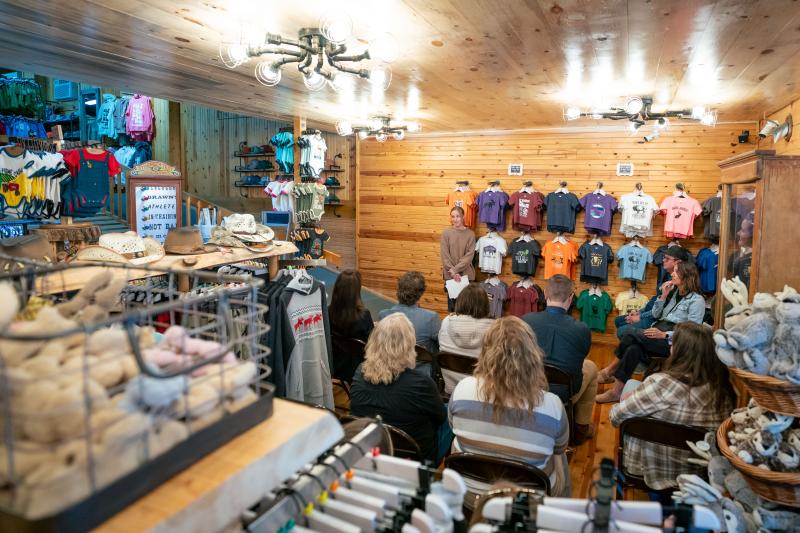 A woman stands by a rack of T-shirts talking to people seated
