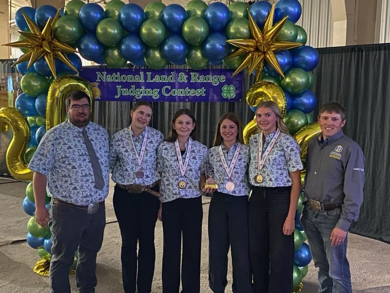 Six people smile in front of a balloon arch