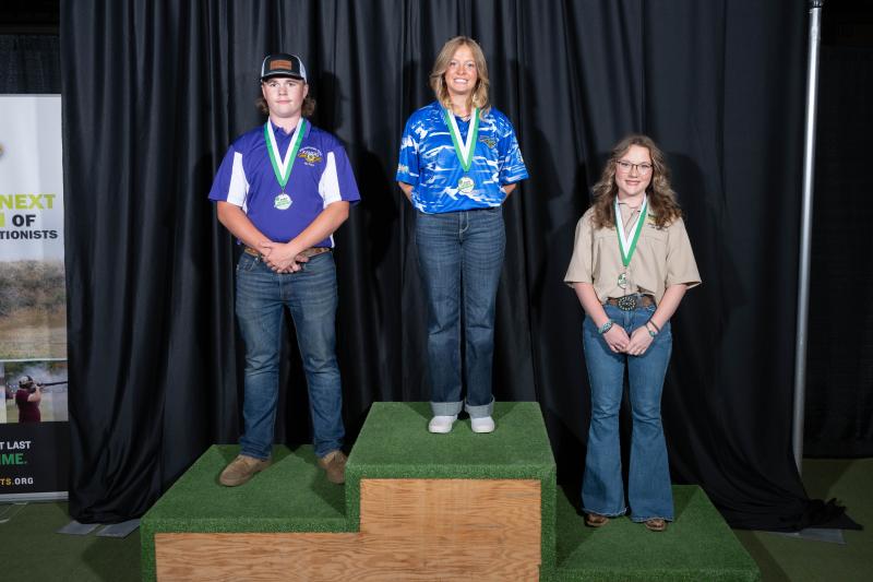 Three youth stand on a podium. A teenage girl in a blue South Dakota shirt stands in the middle with a first-place medal