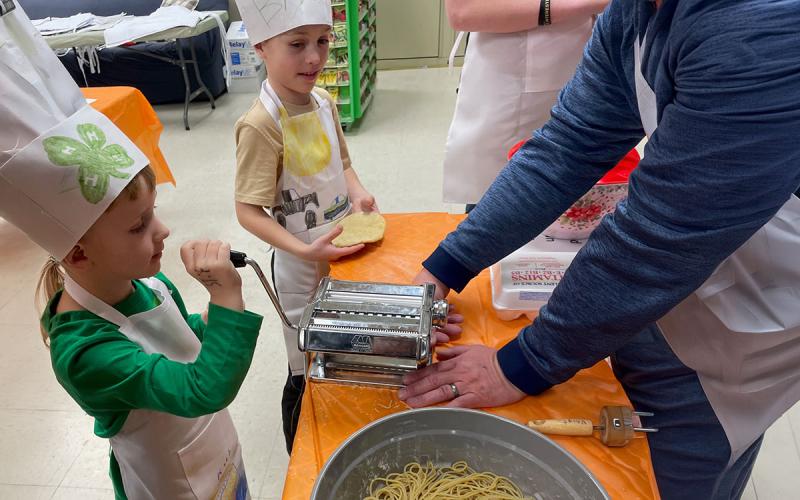 Children making fresh pasta in a kitchen supervised by two adults.