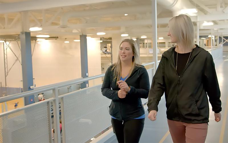 Two young women walking on an indoor track above an athletics facility.