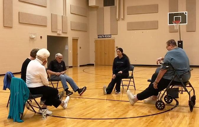 Group of older adults doing a guided foot stretch in a gymnasium.
