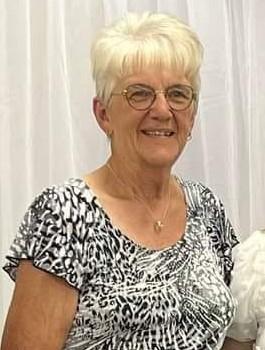 An older woman with short, cropped white hair, wearing glasses and a white-and-black patterned shirt smiles for the camera