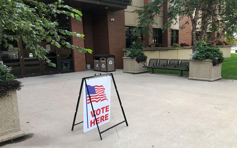 An a-frame sign reading “Vote Here” on the sidewalk outside a local government building.