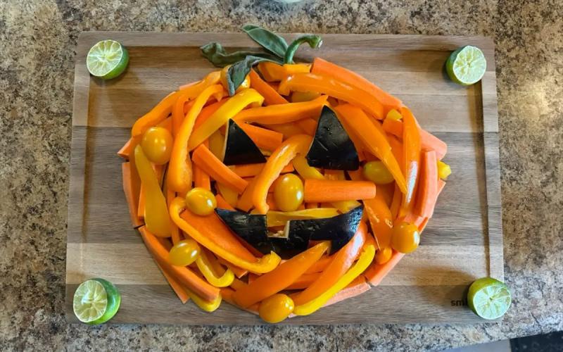 Cut carrots, tomatoes, and eggplant arranged in the shape of a jack-o'-lantern on a cutting board.