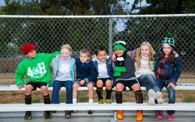 Diverse group of school-age children sitting on bleachers outside in 4-H clothing.; Photos; 4-H Grows Here; Brand; 4-H youth